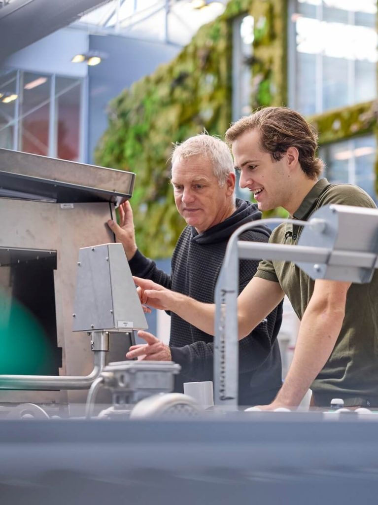 Twee mannen werken samen aan machine in fabriekshal.